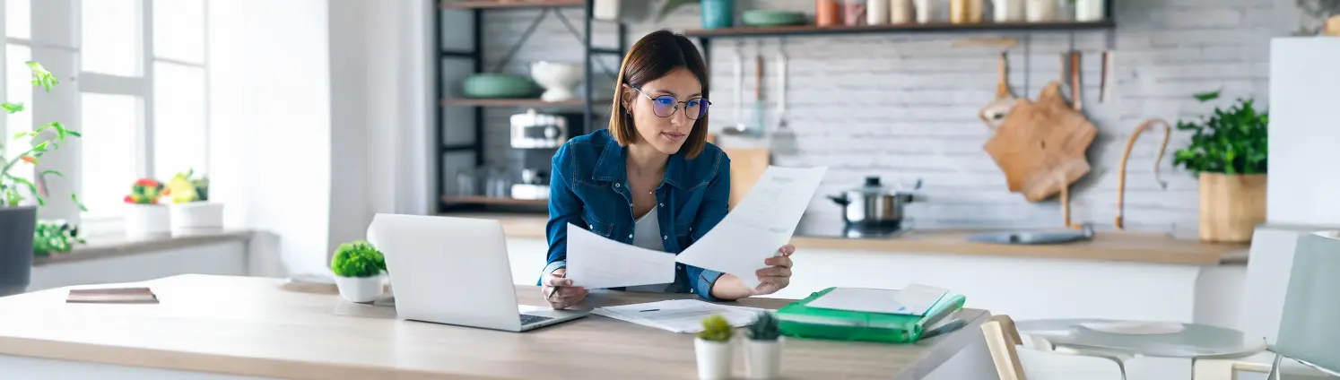 Eine Frau mit Brille sieht sich zwei Seiten Papier an, sie sitzt an einem Schreibtisch mit Laptop