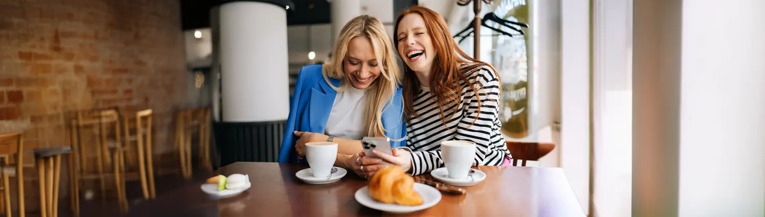 Zwei Frauen sitzen an einem Tisch mit Gebäck, schauen auf einem Smartphone und lachen