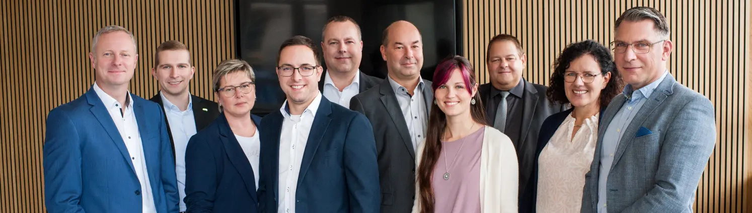 Eine Gruppe aus mehreren Menschen vor einer Holzwand in schicken Outfits schaut in die Kamera