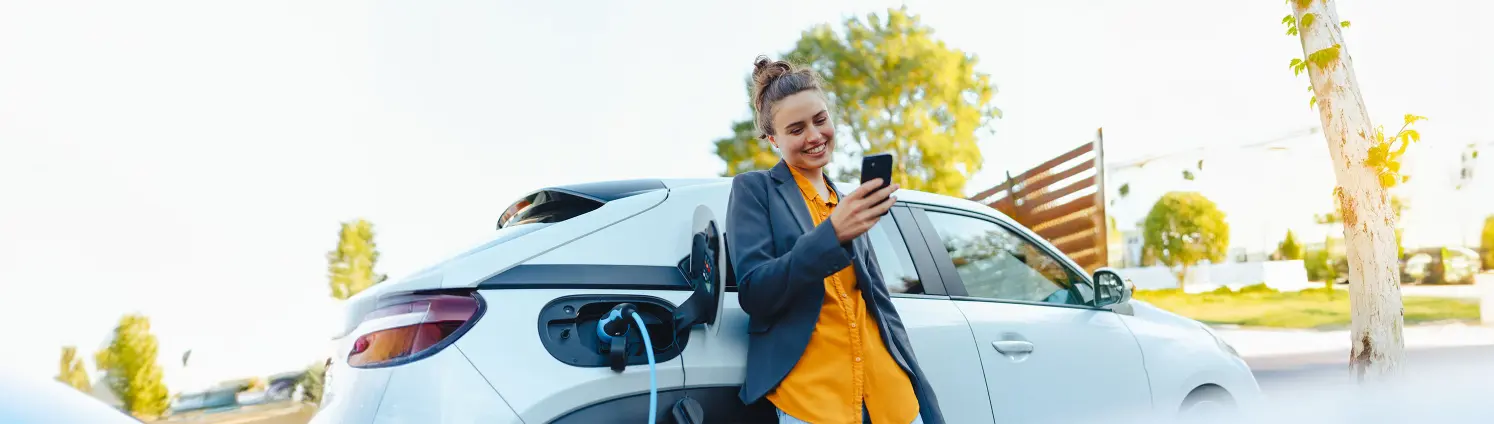 Eine Frau lehnt an einem weißen Auto, das gerade geladen wird und schaut lächelnd auf ihr Handy