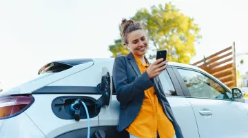 Eine Frau lehnt an einem weißen Auto, das gerade geladen wird und schaut lächelnd auf ihr Handy