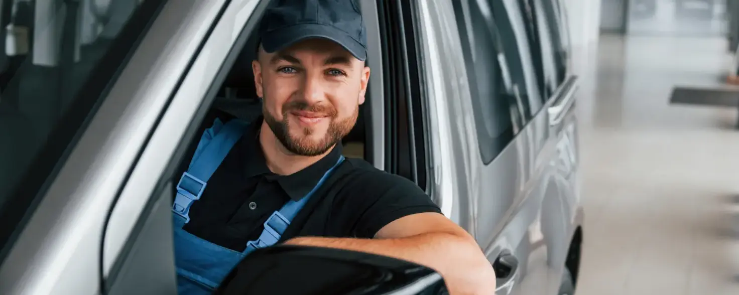 Ein Mann in Monteurkleidung sitz in einem silbernen Auto und lächelt in die Kamera