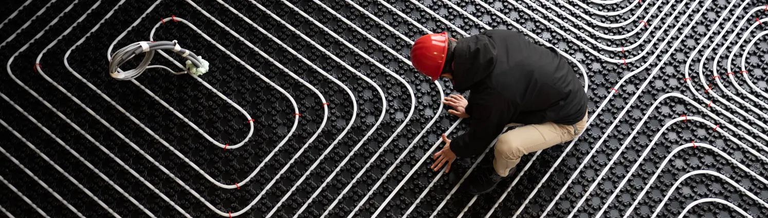 Installateur mit rotem Helm verlegt die Rohre einer modernen Fußbodenheizung.