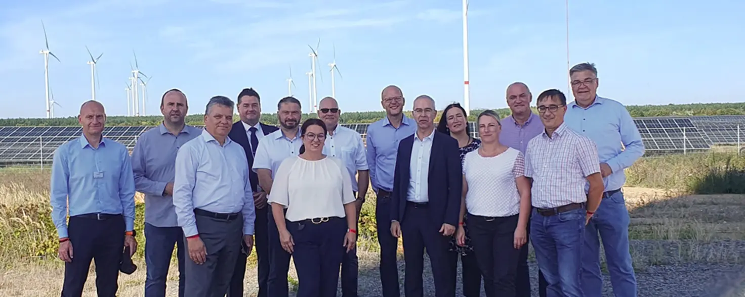 Eine Gruppe aus mehreren Männern und Frauen in schicken Outfits stehen vor einem Photovoltaik-Park und Windkraftanlagen