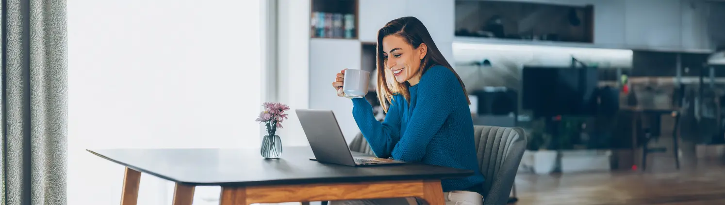 Eine Frau mit Tasse in der Hand schaut lächelnd auf ihren Laptop