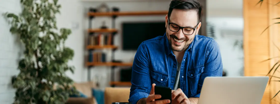 Mann sitzt am Tisch mit Laptop und schaut auf einem Smartphone