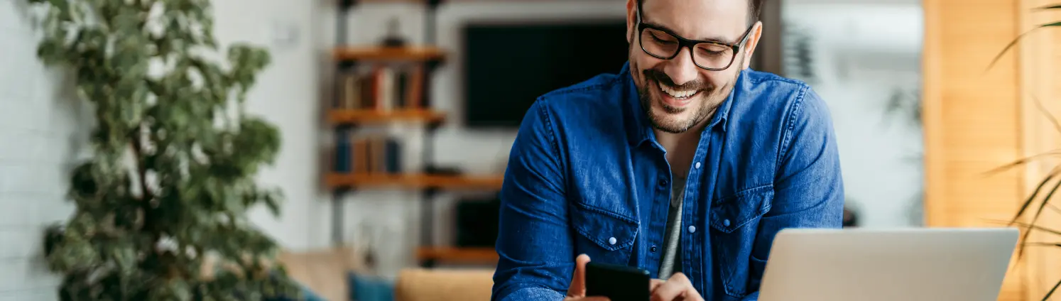 Eine Mann mit Brille sitzt lächelnd vor seinem Laptop und bedient eine Smartphone