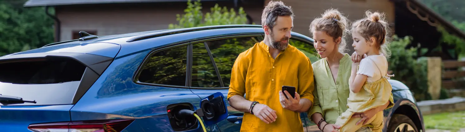 Familie schaut auf einem Smartphone, Hintergrund zeigt ein Haus und ein E-Auto das geladen wird