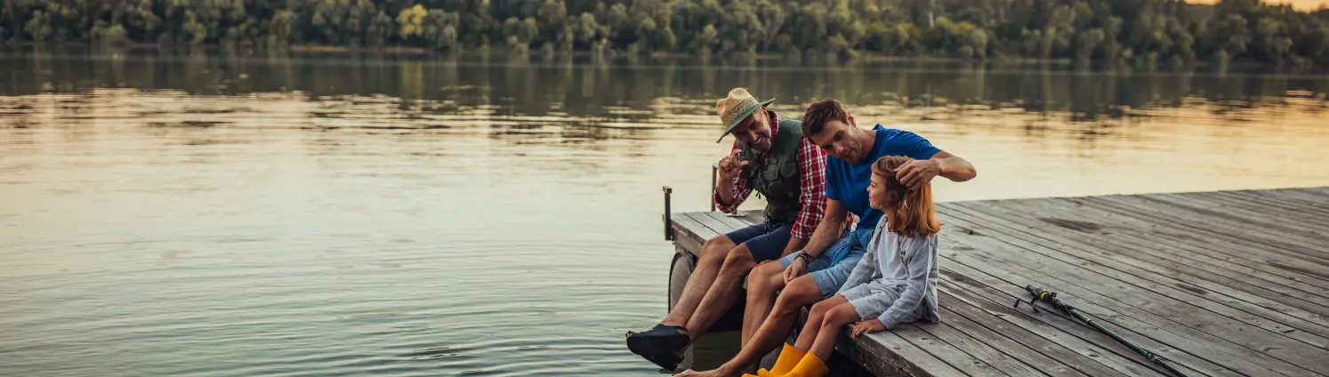 Drei Personen sitzen an einem See auf dem Steg und lassen die Beine baumeln