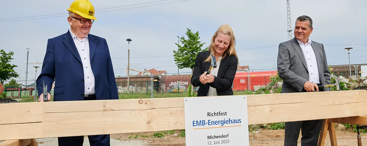 Drei Personen stehen für ein Richtfest auf einer Baustellen vor einem Holzbalken